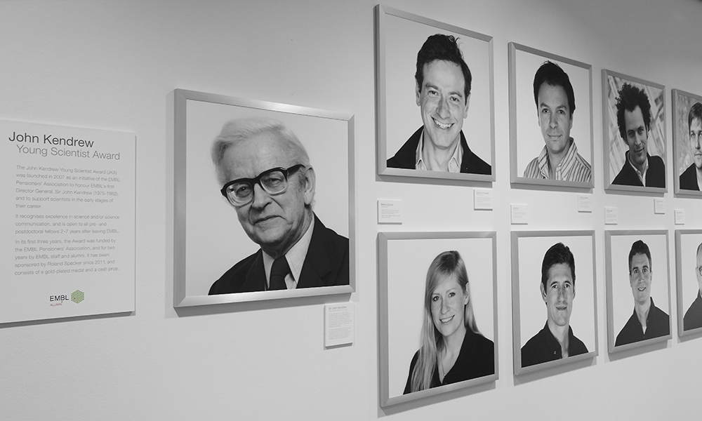Portrait of John Kendrew in front of a line of other portraits in a gallery