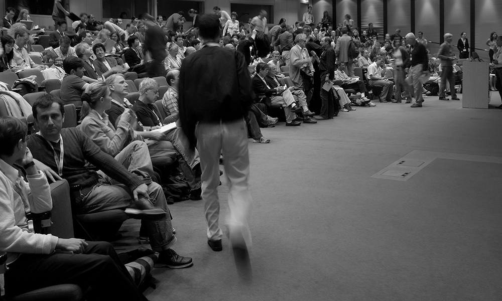 Black and white photo shows a man is walking in a room overfilled with conference participants