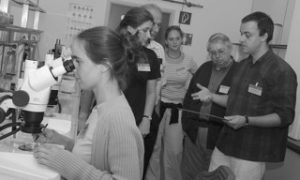 A woman looks into a microscope, while a group of people stand in the background.