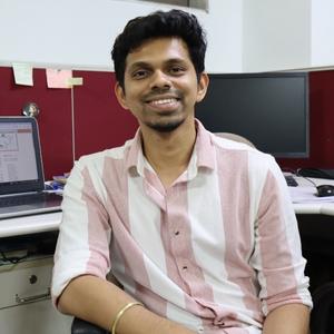 a smiling young man wearing a light coloured striped shirt, sitting in an office chair, with computer screens in the background