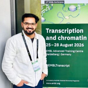 A smiling young man standing in front of a big poster for the 'Transcription and chromatin' conference on the door of the conference venue.