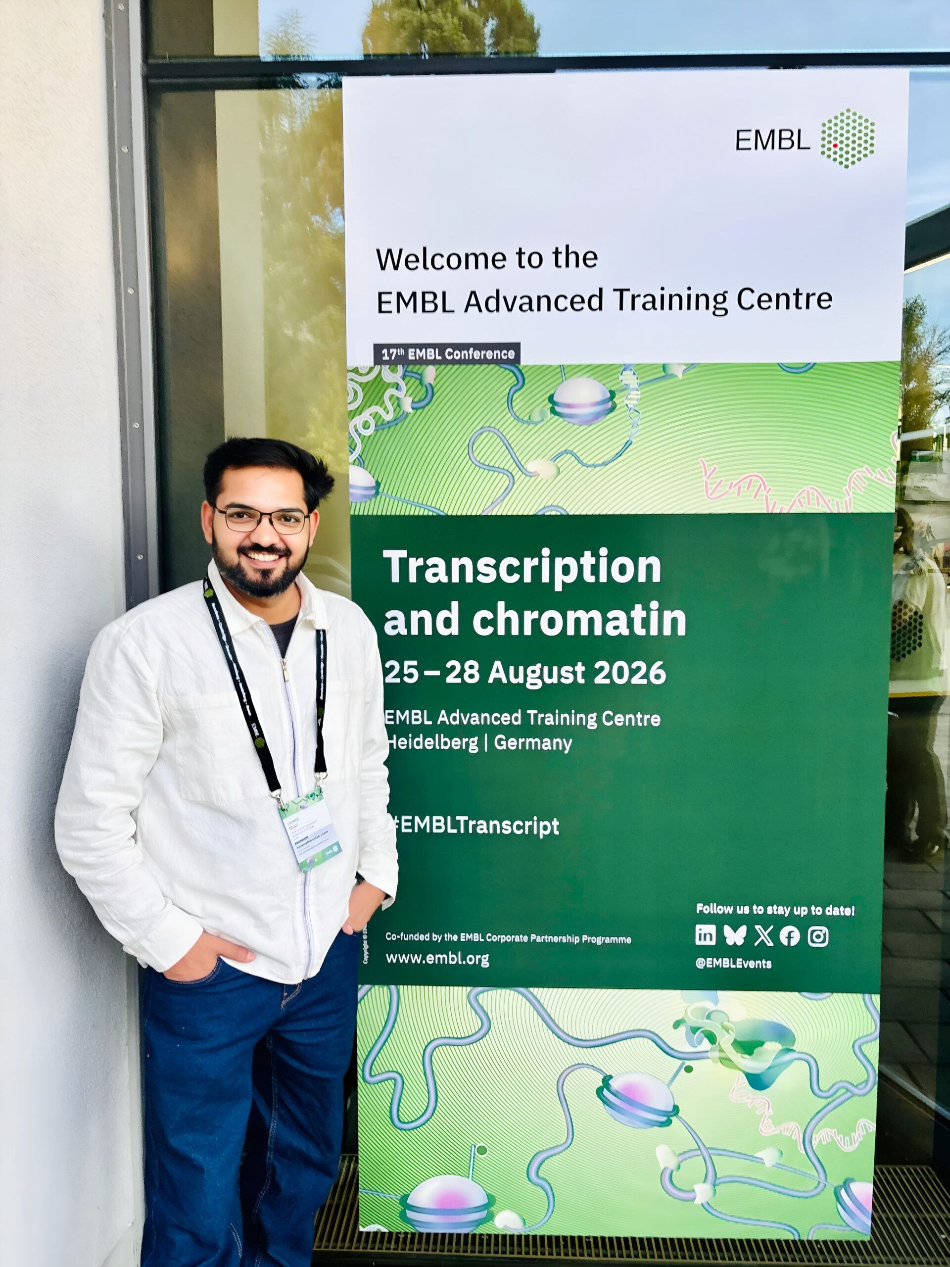 A smiling young man standing in front of a big poster for the 'Transcription and chromatin' conference on the door of the conference venue.