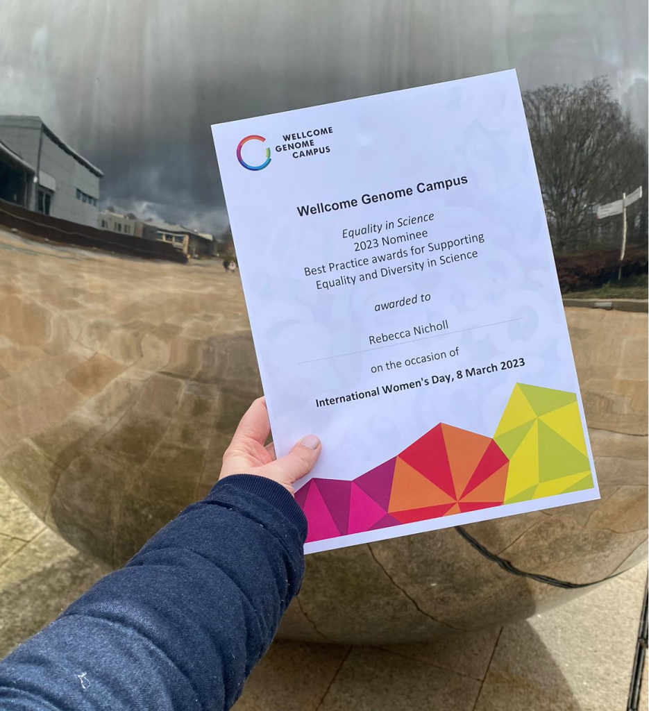 A certificate is held up in front of a large mirrored ball on the grounds of the Wellcome Genome Campus,