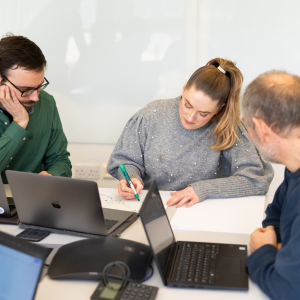 Three people gather around laptops and a sheet of paper taking notes