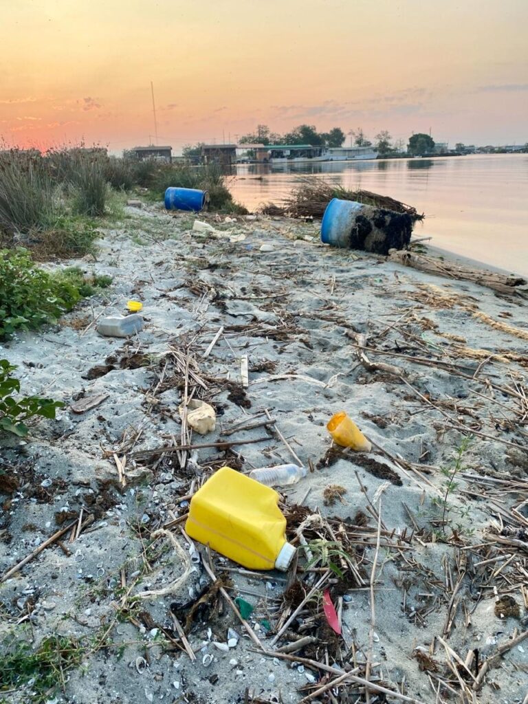 Litter strewn on a beach including a yellow plastic cannister and some rotting blue plastic containers. Sun setting in the background