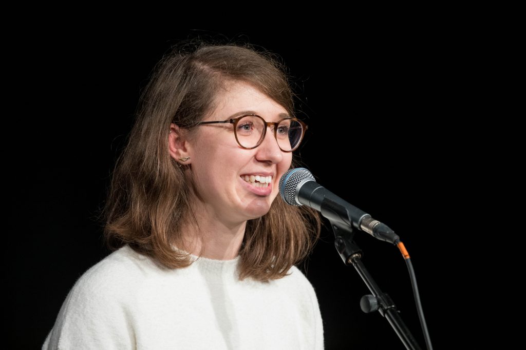 Virginie Uhlmann speaking at Story Collider in Cambridge, she stands in front of a microphone, the backdrop is dark, she is smiling. 