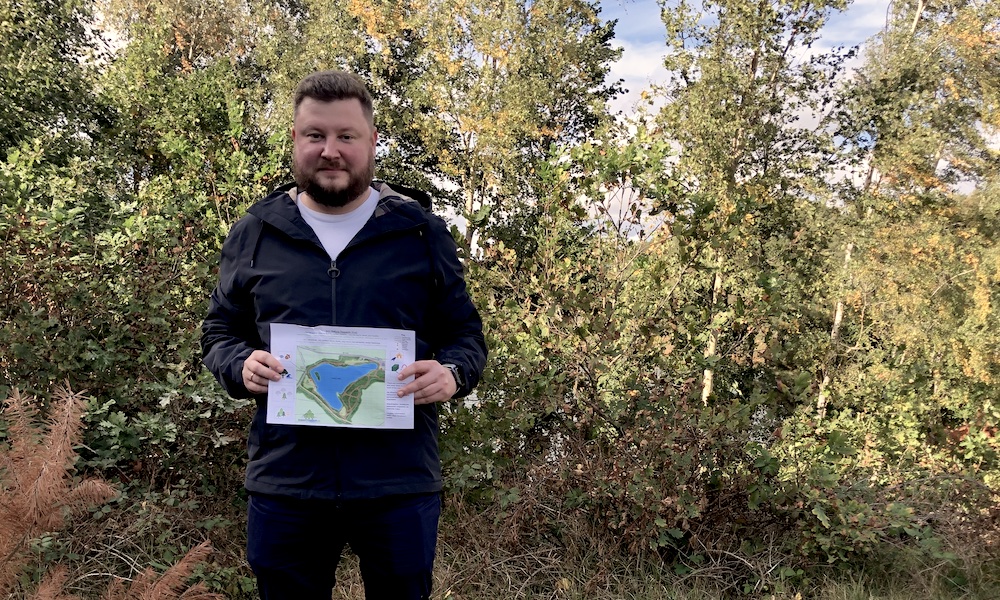 Alexey stands in front of some trees,it's sunny and he is holding the A4 Into Nature map.