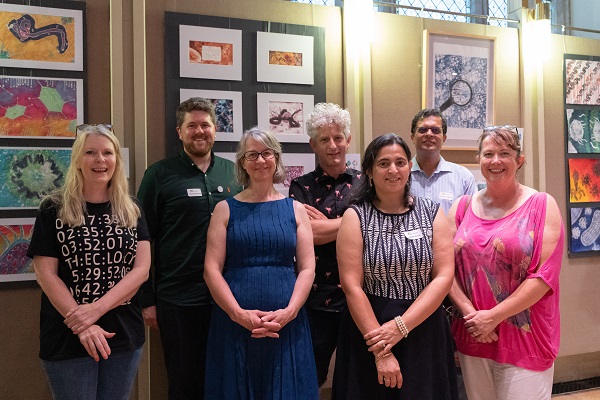 Group of 7 adults pose as a group in front of a wall of artworks from the PDB Art project.