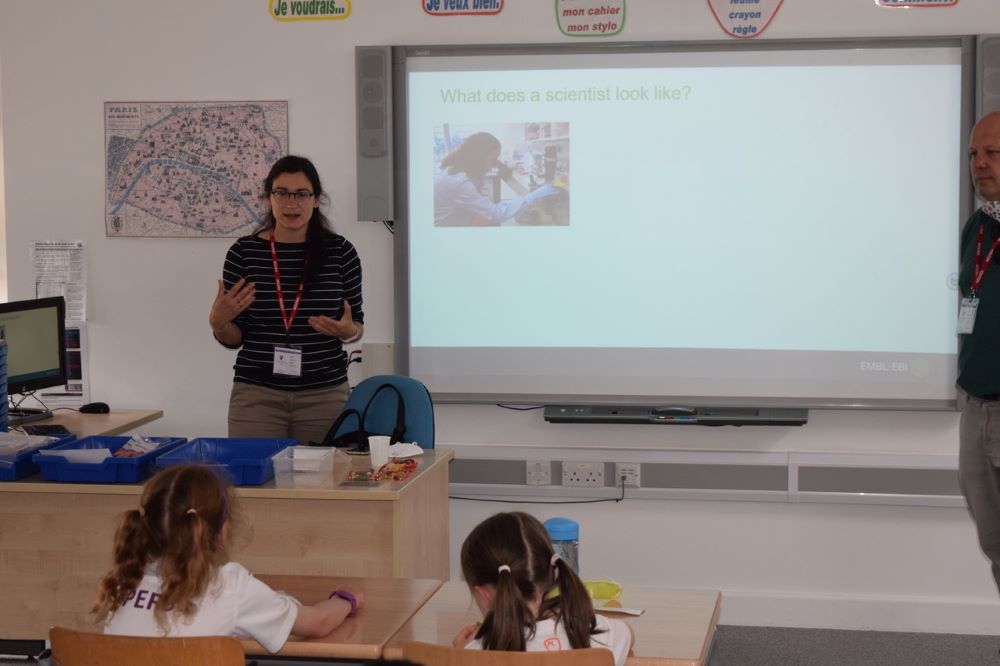 Irene stands in front of a school class talking to the pupils, a powerpoint slide is displayed on a intereactive whiteboard behind her - two pupils in the front row are visible.