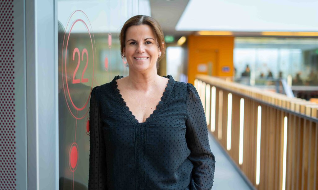 Woman with auburn hair, waring dark top leaning against a glass wall and smiling. She is standing at the end of a corridor.