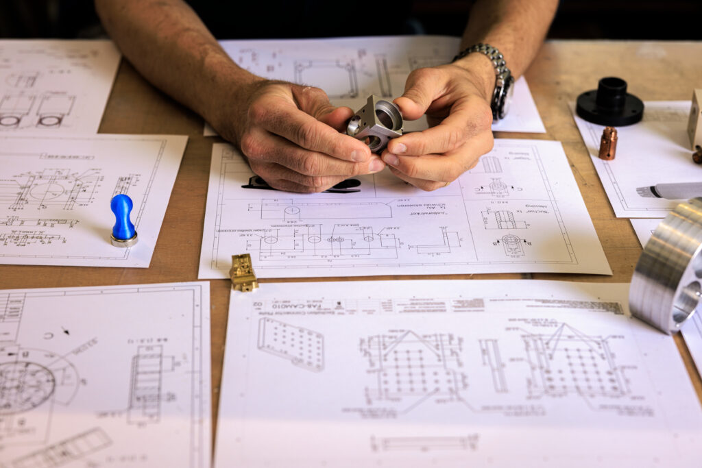 Two hands holding a machined metal part on a bench full of mechanical design drawings