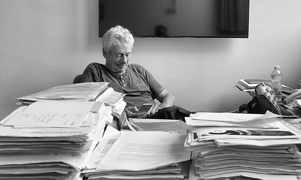 Black and white photo of Phil Avner sitting in his office and looking at his smartphone. a large stack of papers on his desk