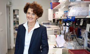 EMBL Director General Edith Heard standing at a laboratory work bench, facing the camera. Edith Heard on the left side, lab bench with pipettes and equipment on the right. Photo taken by érôme Brébion at Institut Curie, Paris.