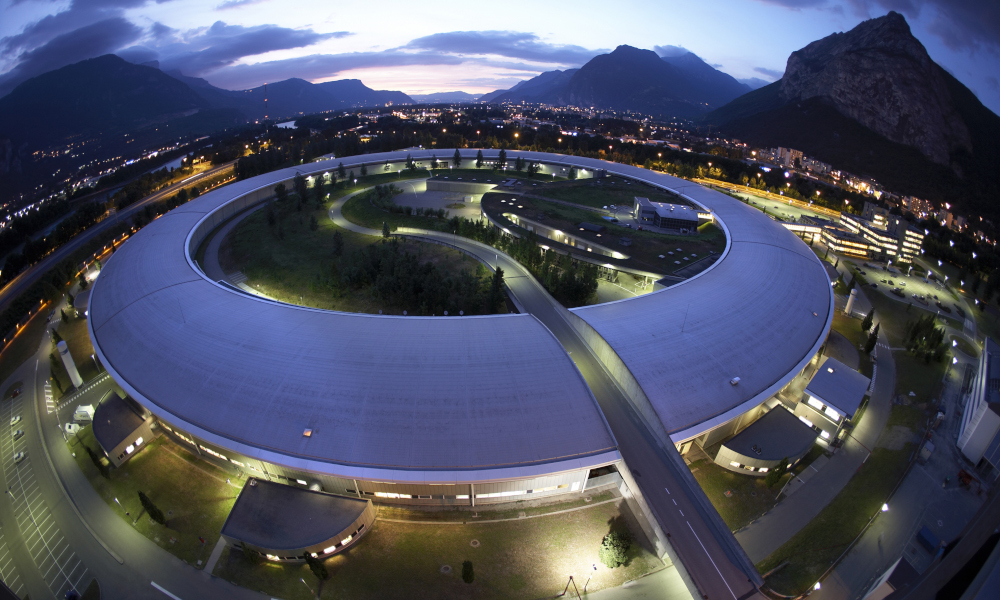 Circular beamline building, surrounded by illuminated Grenoble city in the evening and the alps in the background.