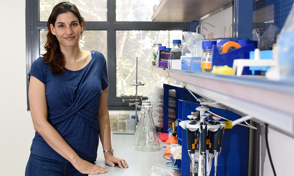 Meytal Landau is standing next to a lab bench. On the bench, laboratory equipment is visible, such as pipettes, flasks, bottles with reagents, a calculator, and more.