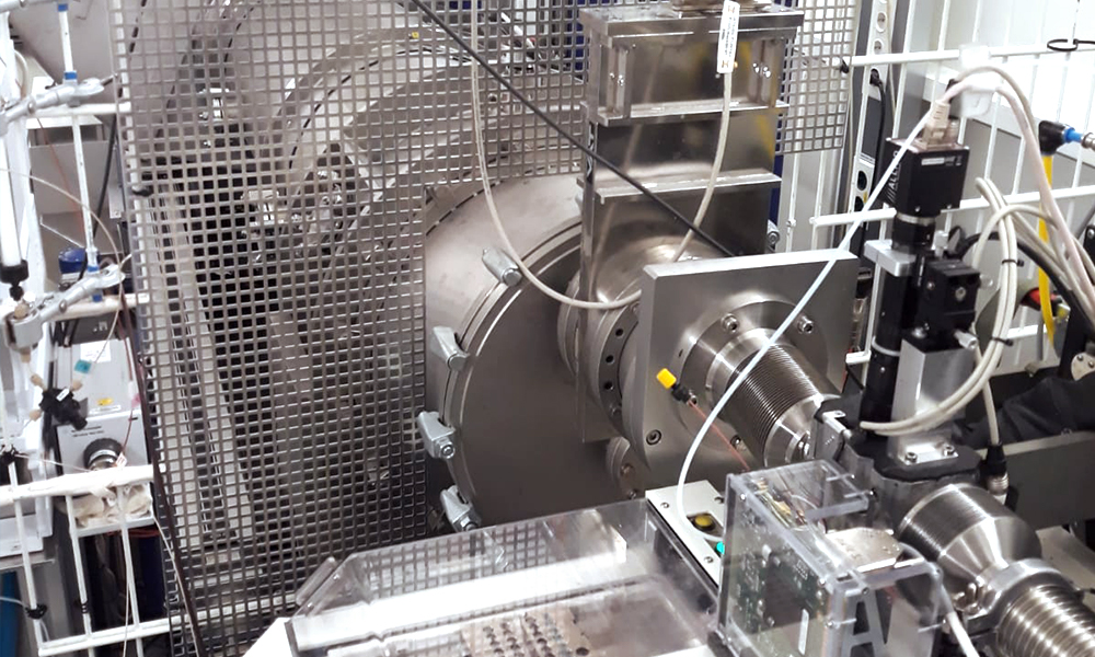 The image shows the beamline P12 at EMBL Hamburg. In the centre, several cylindrical elements are connected into a pipe-like structure. In the front, it is connected to a white box-shaped device, and several smaller devices and cables visible around. There is also a grid visible around the beamline.