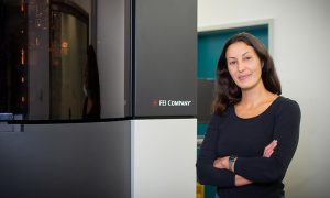 Female scientist stands in front of electron microscope that is taller than she is