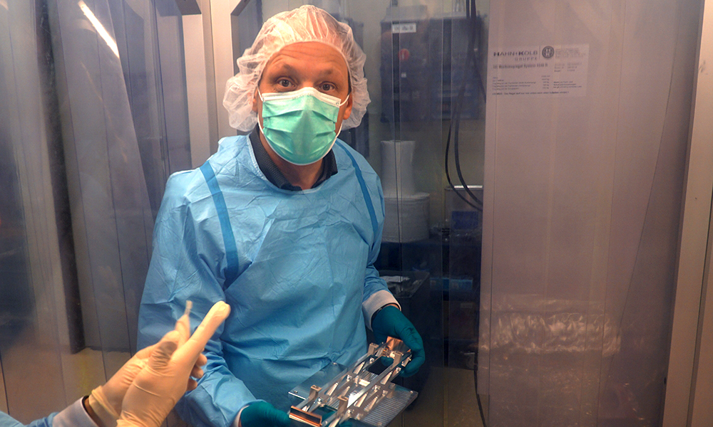 Two scientists dressed in blue coats, wearing gloves, face masks, and hair covers. One of them is holding a metal device that is the mirror support. They are standing next to a table covered with aluminium foil. Around them hang transparent protective sheets that are part of a laminar flow tent, allowing them to work under clean-room conditions.