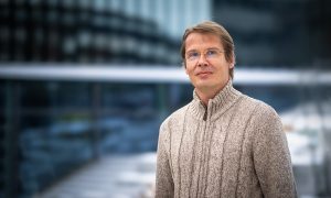 man wearing sweater and glasses stands in front of snow and glassy building