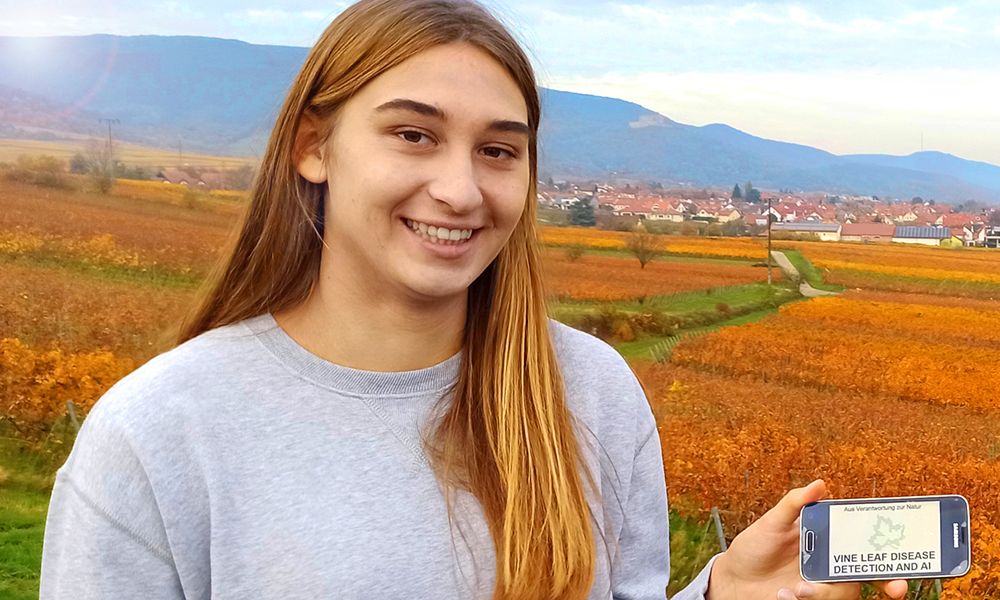 Maria-Theresa Licka holding a smartphone displaying an app she developed. Vineyards, houses and hills in the background.