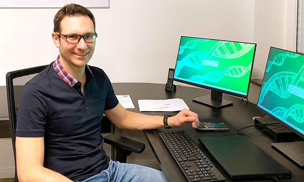 Man in black t-shirt and jeans wearing glasses sits in front of two computer screens.