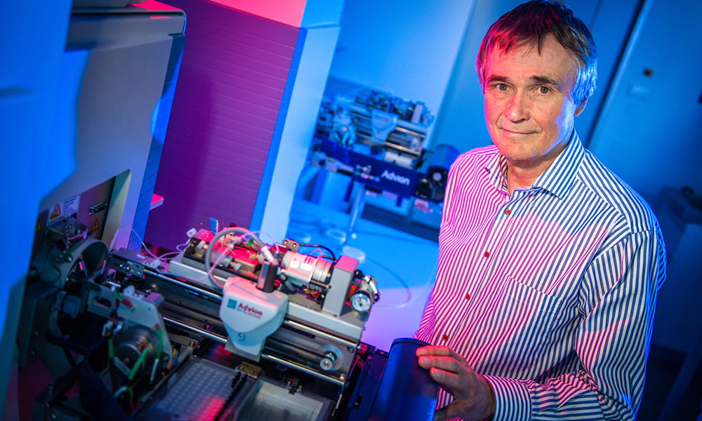 Older male scientist stands in front of machinery in a blue-lit room
