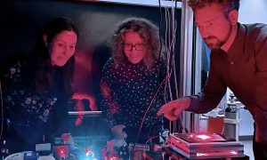 Three researchers surround microscopy equipment in a dark room with red lighting.