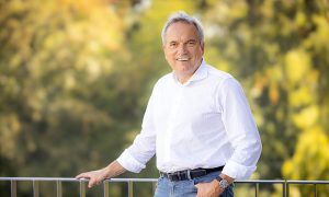 Man in white shirt and blue jeans standing on a terrace in front of trees, facing the camera.