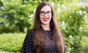 woman with long brown hair is smiling in front of shrubbery outside