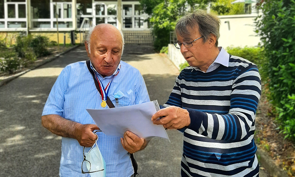 Two men stand outside in front of a building, holding a document and looking at it.