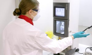 Female scientist with lab coat, gloves, safety goggles, and mask holds a pipette to inject a protein sample into a machine.