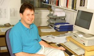 Male person sitting at a desk with computer and papers, looking at the camera.