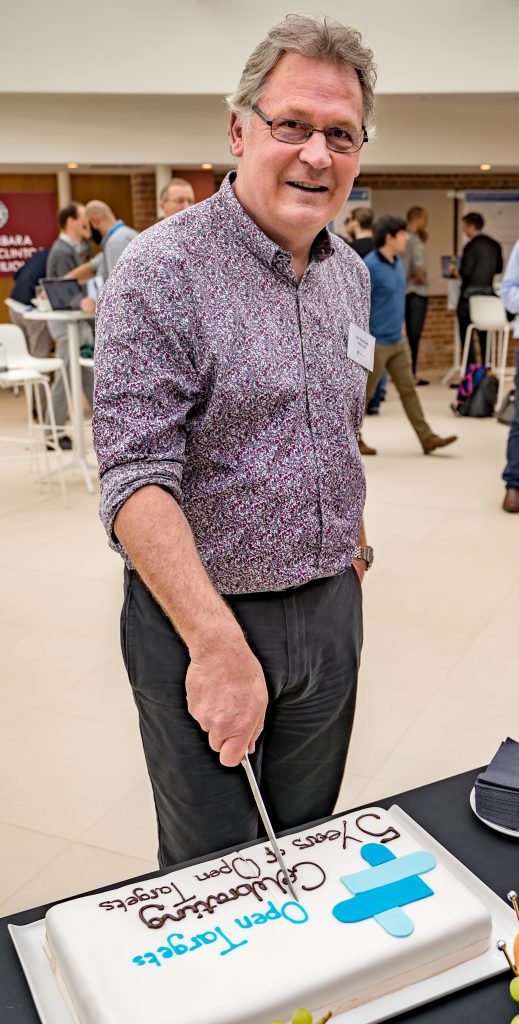 Male person standing behind a table, looking at the camera, holding a knife to cut a cake.
