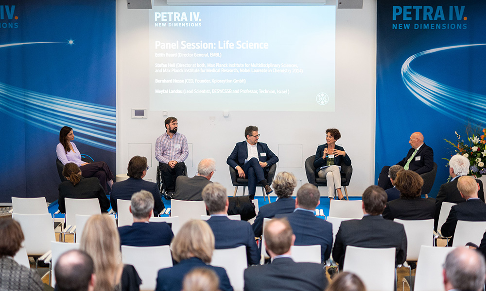 Photo of the speakers during the panel discussion. Edith Heard is talking. In front of the panel, there is the audience seen from the back, and behind the panel there are banners with the DESY PETAR IV logos and a slide displaying the names of the panellists.