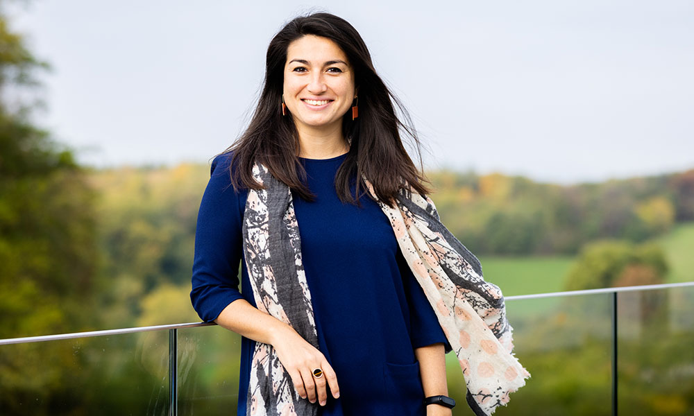 A female scientist in a blue dress stands in front of blurred woodsy background