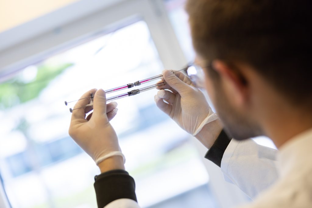 Over shoulder photo showing Kirill Kovelev raising two syringes with pink and purple liquid inside to examine them against the light from a window.
