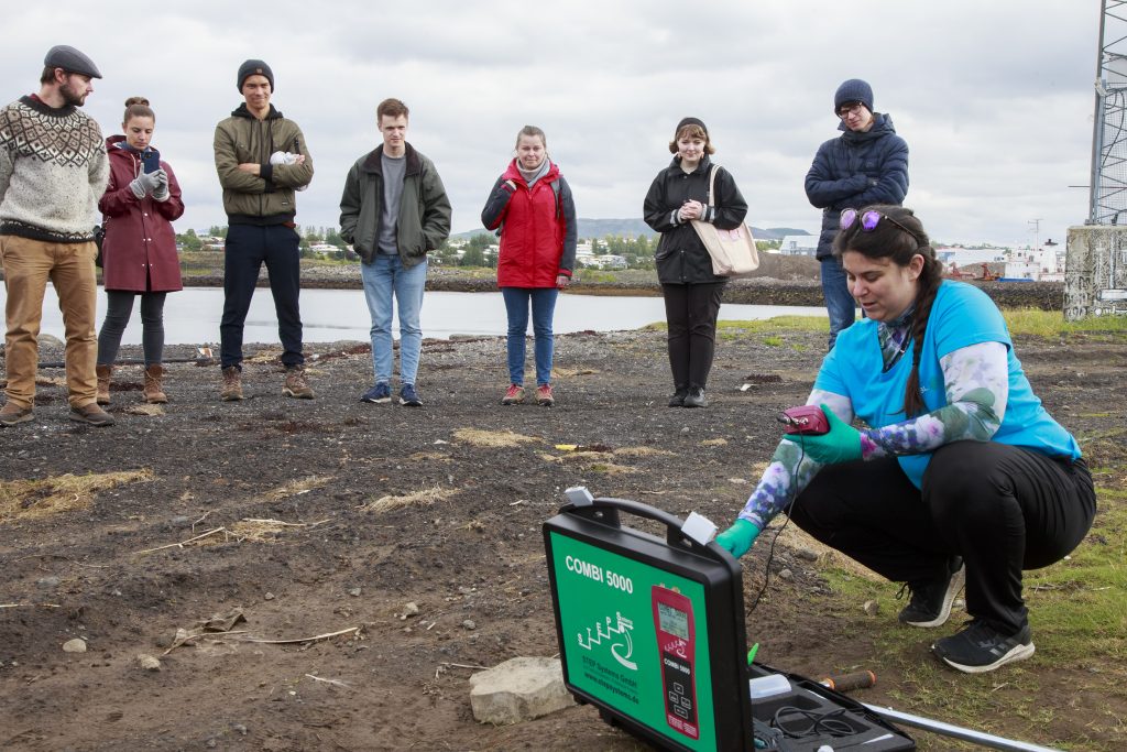 A female scientist demonstrates something with a machine on a beach while onlookers look on. 