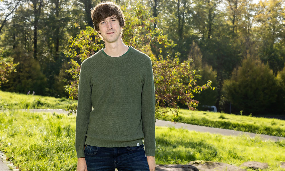 male scientist in green shirt and dark pants stands outside in front of tree