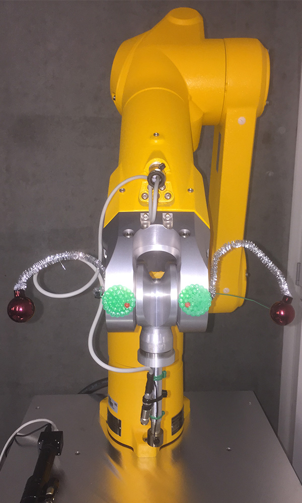 Yellow robotic arm disguised as a bee. Two EMBL logos on the sides resemble compound eyes, two shiny wires with Christmas baubles above the ‘eyes’ look antennae, and the gripper is the mouth part. 