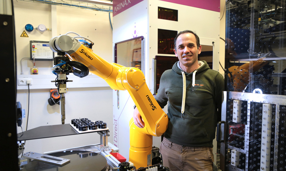 Moisés Bueno is standing next to the transfer robot. The robot has a form of a yellow robotic arm on a stand. Behind is the CrystalDirect™ Harvester, which is a white cuboid with two transparent dimmed windows for laser protection.