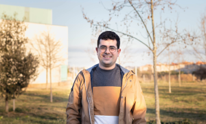 Portrait of a person looking at the camera. In the background there are trees and a white building