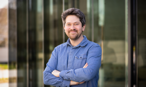 Male scientist in blue shirt standing in front of a building with glass doors.