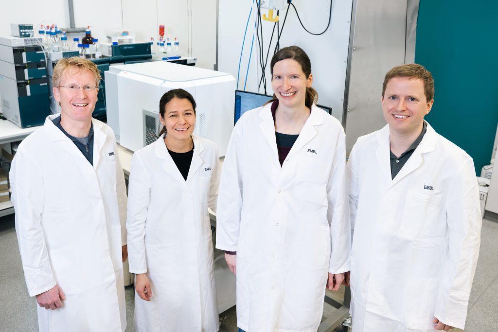 Four scientists wearing lab coats and standing in front of an ICS machine. 