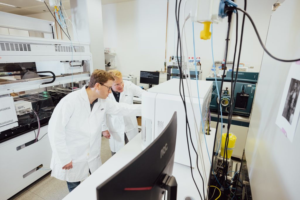 Two male scientists wearing lab coats pointing towards something inside an ICS machine. 