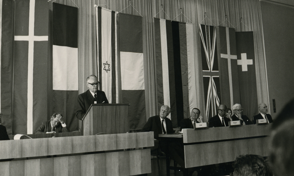 Black and white photo of 7 men in front of country flags