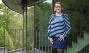 A female scientist wearing a blue skirt and striped blue shirt stands outside.