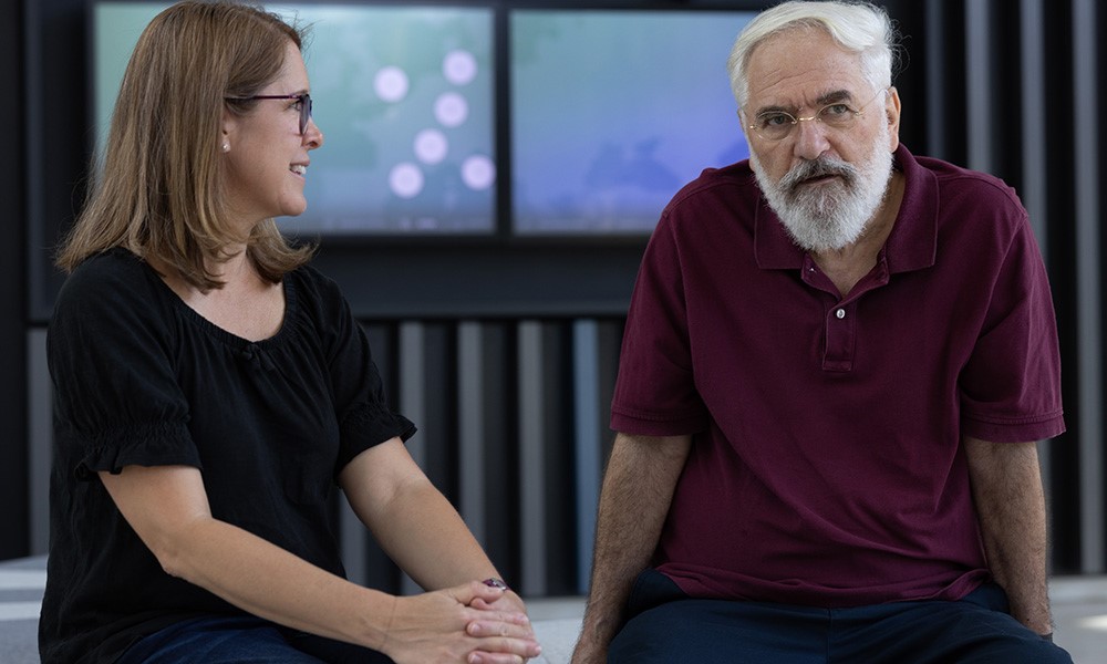 A female scientist with shoulder-length hair and black shirt listens to a male scientist in burgundy shirt and white beard, seated to the right.