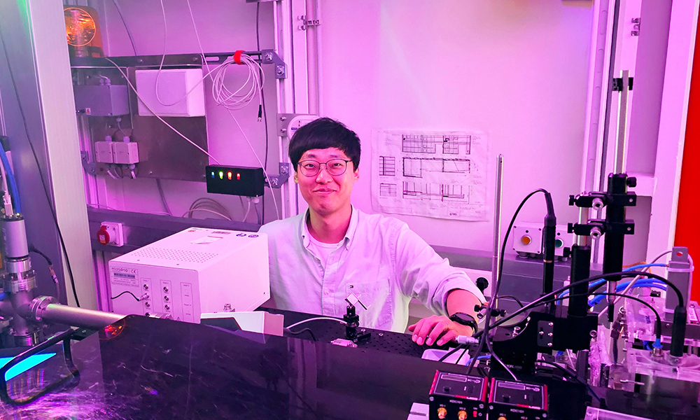 Sihyun Sung is standing next to experimental equipment for time-resolved serial X-ray crystallography. The light in the room is pink.