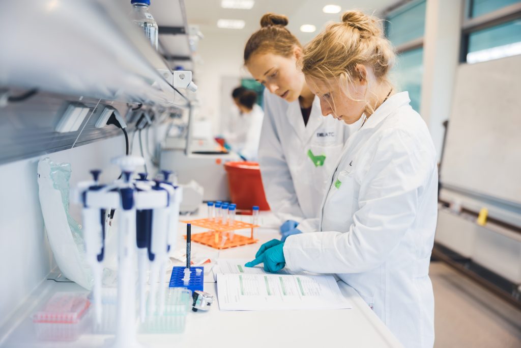 Two female scientists in a lab, wearing lab coats and looking down at a handout
