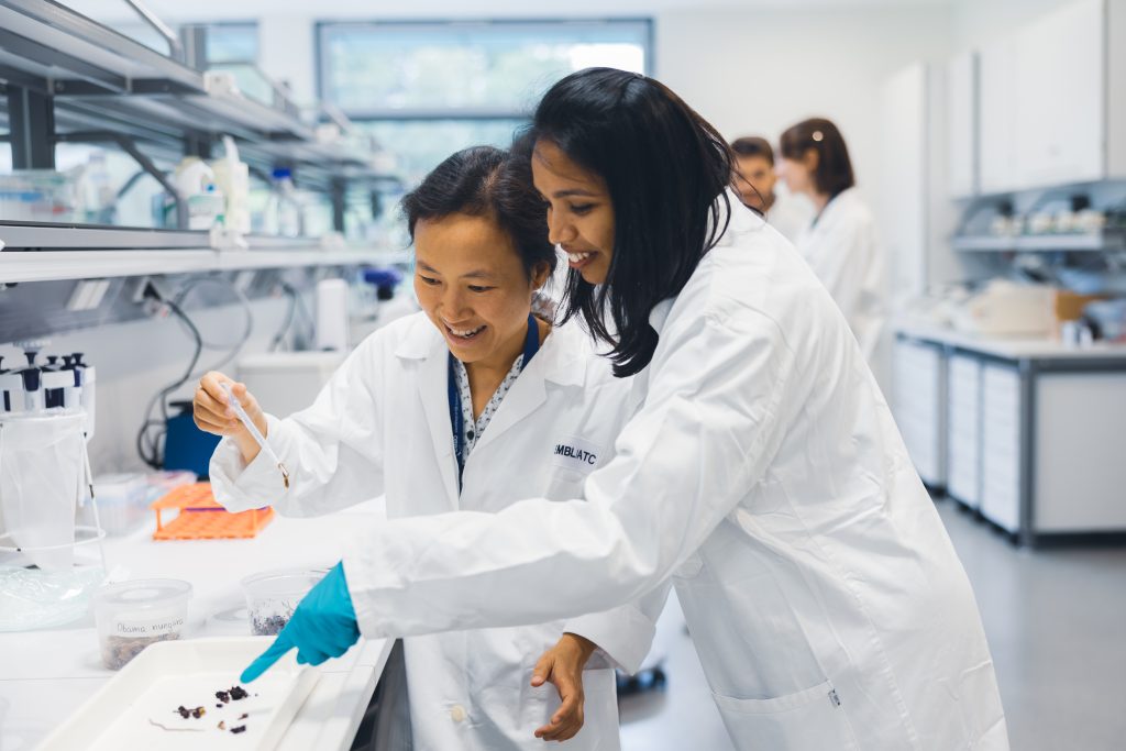 Two female scientists in lab coats, one holding a flatworm using a dropper, another pointing at the sample. 
